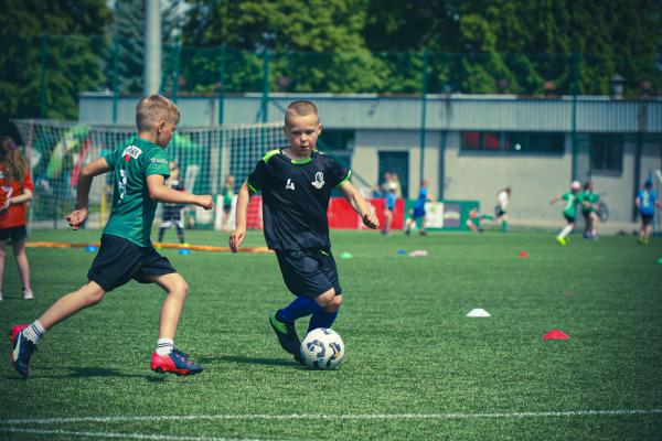 Dwaj chłopcy na zielonym boisku: chłopiec w granatowej koszulce prowadzi piłkę, a obok w zielonym stroju biegnie drugi zawodnik.