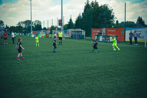 Podczas finału wojewódzkiego Tymbarka dzieci w czarnych i neonowożółtych strojach walczą o piłkę na zielonym boisku, a w tle stoi sędzia.