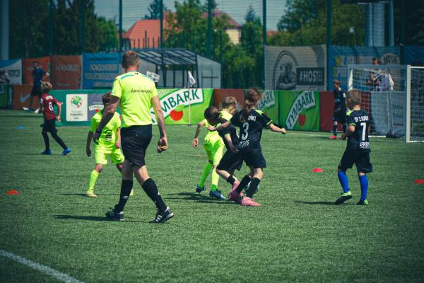 Mali piłkarze w czarnych i neonowo-żółtych strojach walczą o piłkę na zielonym boisku, obok idzie sędzia w neonowej koszulce.