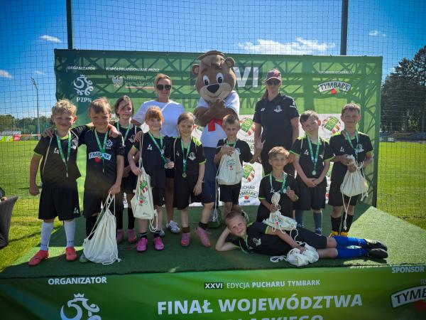 Dzieci w czarnych strojach z zielonymi akcentami, z medalami na szyjach, pozują na zielonym podium z maskotką niedźwiedzia i dwiema dorosłymi kobietami.