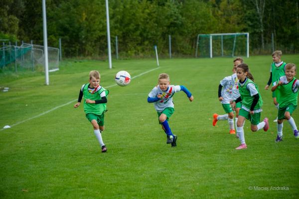 Młodzi piłkarze w wieku U9 biegną za piłką na zielonym boisku; część ma zielone znaczniki, reszta białe koszulki, w tle bramka i drzewa.