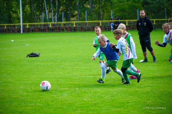 Żacy U9 walczą o piłkę na zielonym boisku; chłopiec w niebieskiej kamizelce prowadzi piłkę, obok zawodnicy w białych koszulkach.
