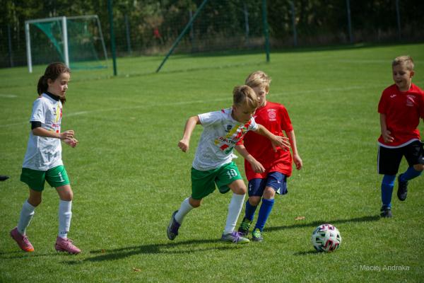 Chłopiec w białej koszulce i zielonych spodenkach prowadzi piłkę na boisku, obok biegną dzieci w czerwonych strojach.