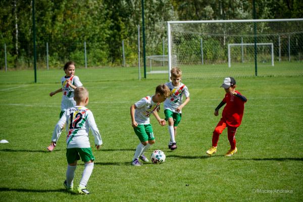 Mali piłkarze U9 w białych koszulkach i zielonych spodenkach walczą o piłkę na boisku, a po prawej stoi chłopiec w czerwonym dresie i czapce.