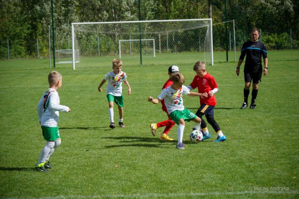 Chłopcy z kategorii U9 walczą o piłkę na boisku: kilku w biało-zielonych strojach, jeden w czerwonym; z boku sędzia w czarnym stroju.