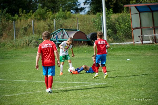Chłopcy w czerwonych koszulkach i zawodnik w białej koszulce z zielonymi spodenkami walczą o piłkę na trawie; jeden z nich siedzi na ziemi.