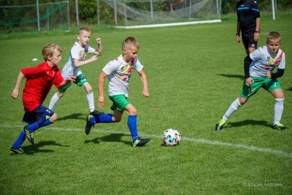 Dzieci z kategorii żak U9 biegną za piłką na zielonym boisku: chłopiec w czerwonej koszulce i trzech w białych koszulkach z zielonymi spodenkami.