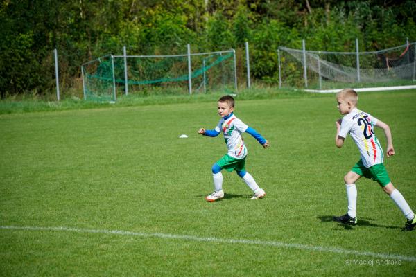 Dwaj chłopcy w białych koszulkach z kolorowymi pasami i zielonych spodenkach biegną po trawiastym boisku; po prawej zawodnik z numerem 25 na plecach,…