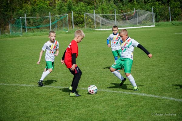 Czterech chłopców na boisku: dwóch w białych koszulkach z zielonymi spodenkami grających w piłkę, obok zawodnik w czerwonej koszulce, a z tyłu kolejny…