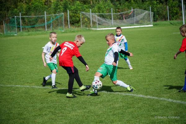 Dwaj chłopcy, jeden w czerwonej koszulce z numerem 9, drugi w białej z zielonymi spodenkami, walczą o piłkę na boisku podczas meczu żaków U9.