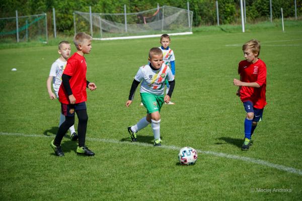 Mecz żaków U9: chłopiec w białej koszulce i zielonych spodenkach prowadzi piłkę, obok biegną zawodnicy w czerwonych strojach.