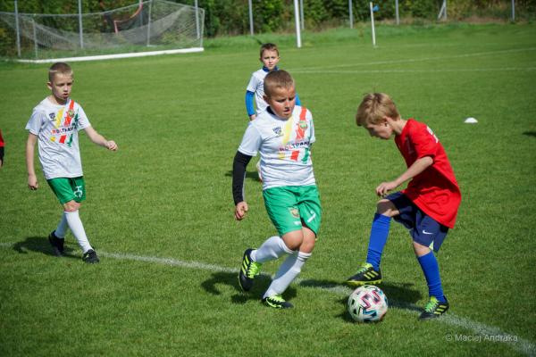 Dzieci w białych koszulkach z pasami i zielonych spodenkach prowadzą piłkę na trawiastym boisku, a chłopiec w czerwonej koszulce próbuje ją odebrać. L…
