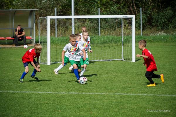 Dzieci U9 walczą o piłkę na boisku: chłopiec w białej koszulce prowadzi ją, a dwaj zawodnicy w czerwonych strojach próbują go zatrzymać.