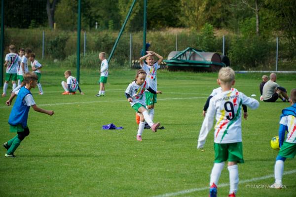 Dzieci w białych koszulkach z kolorowymi pasami i zielonych spodenkach na boisku; z prawej chłopiec trzyma żółtą piłkę.