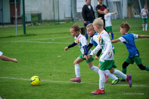 Dzieci U9 biegną za żółtą piłką po murawie; dziewczynka z kucykiem prowadzi akcję, obok chłopcy w białych koszulkach i niebieskich znacznikach.