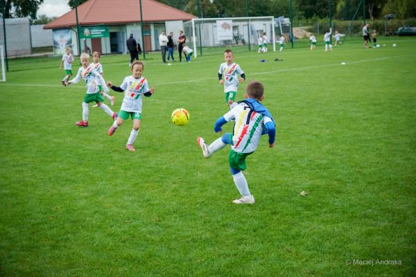 Podczas meczu żaków U9 chłopiec w białej koszulce w kolorowe pasy uderza żółtą piłkę, a obok biegną rówieśnicy w zielonych spodenkach.