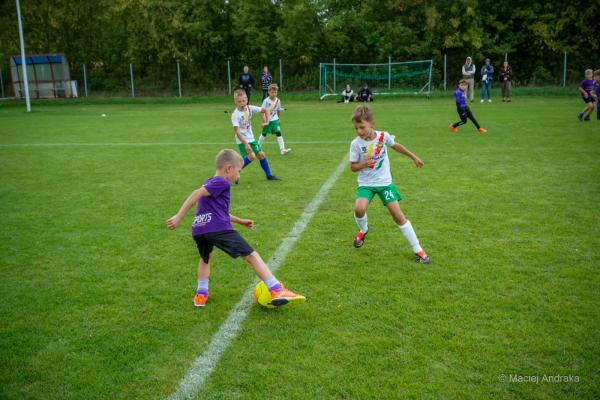 Chłopiec w fioletowej koszulce kopie piłkę przy białej linii, obok biegnie rywal w białej koszulce i zielonych spodenkach.