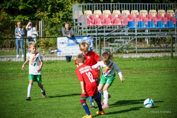Mecz żaków U9 na zielonej murawie: dziewczynka w białym stroju prowadzi piłkę, obok biegną chłopcy w białych i czerwonych koszulkach.