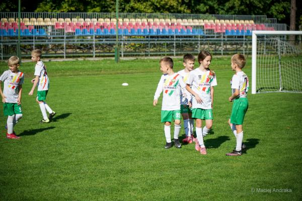 Grupka dzieci w białych koszulkach z kolorowymi pasami i zielonych spodenkach rozmawia na boisku obok bramki podczas meczu żaków U9.