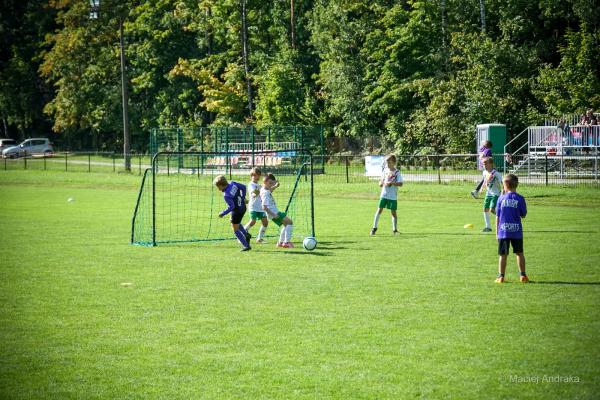 Młodzi piłkarze w fioletowych i białych strojach walczą o piłkę przy bramce na zielonym boisku; w tle drzewa i trybuny.
