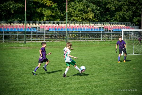 Chłopiec w białym stroju uderza piłkę na zielonej murawie, obok biegnie zawodnik w fioletowej koszulce, a bramkarz czeka przy bramce.