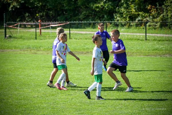 Dzieci z kategorii U9 na zielonym boisku: dwoje w białych koszulkach i zielonych spodenkach oraz kilkoro w fioletowych strojach przy ogrodzeniu.