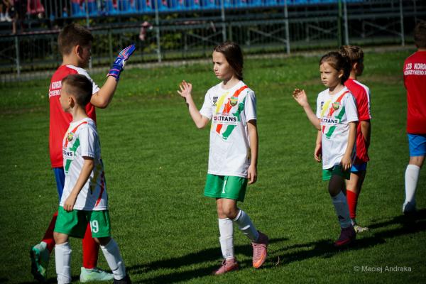 Dzieci na boisku: chłopiec w czerwonym stroju i rękawicach unosi dłoń, obok dziewczynki w białych koszulkach z zielonymi pasami idą na mecz.