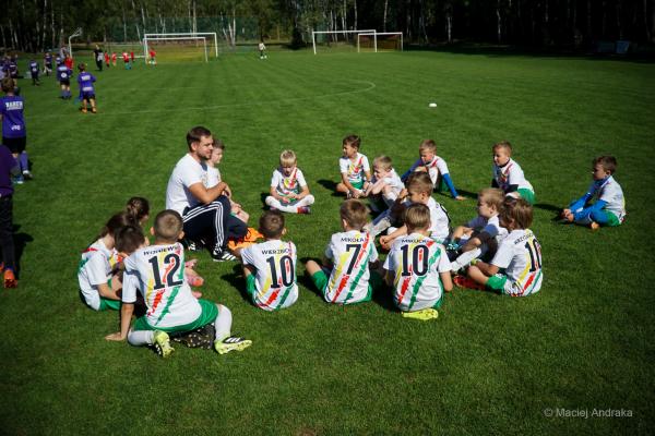 Żacy U9 w białych koszulkach z kolorowymi pasami siedzą na trawie w kręgu wokół mężczyzny w białej koszulce i czarnych spodniach.