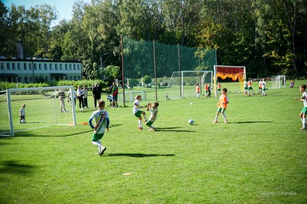 Żacy grają na słonecznym boisku: chłopcy w biało-zielonych koszulkach walczą o piłkę, a obok biegną zawodnicy w pomarańczowych znacznikach.