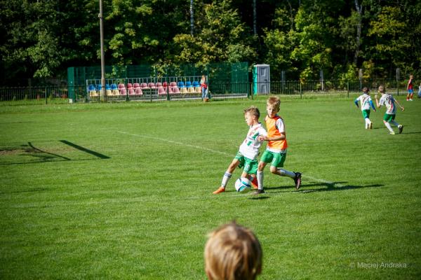Dwóch chłopców w białych koszulkach z zielonymi spodenkami walczy o piłkę na boisku; jeden w pomarańczowej kamizelce treningowej, a w tle kilku zawodn…