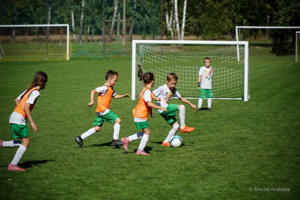 Dzieci w pomarańczowych znacznikach i białych koszulkach z zielonymi spodenkami walczą o piłkę na zielonej murawie przy bramce.