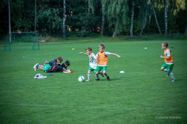 Dzieci U9 na boisku: dwóch chłopców w białych koszulkach prowadzi piłkę, obok biegnie chłopiec w pomarańczowym znaczniku, bramkarz leży na trawie.