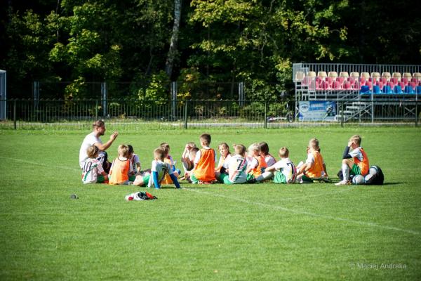 Młodzi piłkarze siedzą na trawie w kręgu wokół trenera, który coś tłumaczy; w tle widać trybuny z czerwonymi i niebieskimi krzesełkami.
