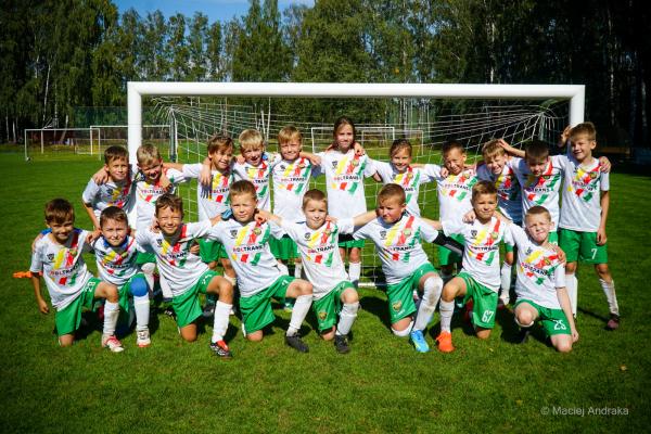 Drużyna żaków U9 w białych koszulkach z kolorowymi pasami i zielonych spodenkach pozuje przy bramce na boisku, w tle drzewa.