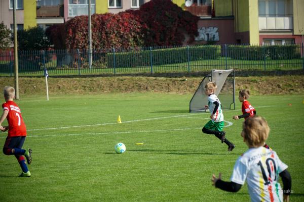 Dzieci z kategorii U9 na zielonym boisku: chłopiec w białej koszulce i zielonych spodenkach biegnie za piłką, obok gracze w czerwonych strojach.