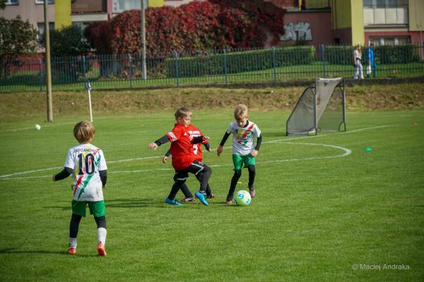 Dwóch chłopców w czerwonych strojach walczy o piłkę z zawodnikiem w białej koszulce i zielonych spodenkach na boisku podczas meczu żaków U9.