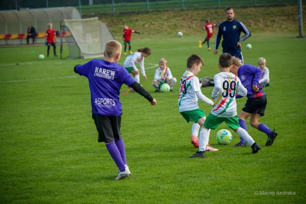 Dzieci w wieku żaka walczą o piłkę na zielonym boisku: dwóch chłopców w biało-zielonych strojach i zawodnik w fioletowej koszulce.