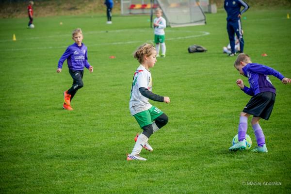 Żacy U9 na zielonym boisku: chłopiec w biało-zielonym stroju biegnie w centrum, a z prawej chłopiec w fioletowych barwach prowadzi piłkę.
