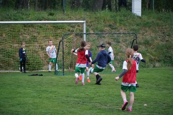 Dzieci w białych koszulkach grają w piłkę na trawiastym boisku; kilka ma czerwone znaczniki, a bramkarz stoi w ciemnym dresie.