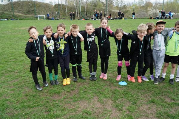 Grupa dzieci w czarnych i granatowych strojach, z medalami na szyjach, stoi w rzędzie obejmując się na zielonym boisku.