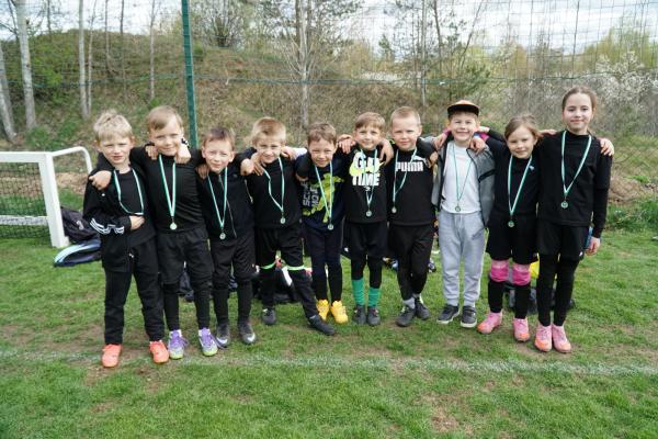 Grupa dziewięciorga dzieci w czarnych strojach sportowych stoi w rzędzie, obejmując się, na szyjach mają zielone medale; w tle siatka ogrodzenia i zielone drzewa.