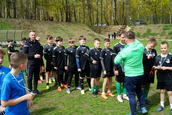 Młodzi piłkarze w czarnych strojach stoją w szeregu na boisku, a mężczyzna w zielonej bluzie do nich mówi; kilku chłopców ma medale na szyi.