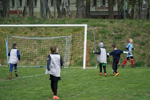 Młodzi piłkarze w białych znacznikach i granatowych koszulkach biegną po zielonym boisku w stronę bramki, a jeden z nich prowadzi piłkę.