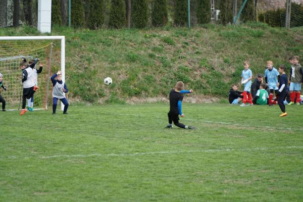 Młody piłkarz w czarnym stroju z niebieskimi rękawami kopie piłkę przy bramce, a obok stoją zawodnicy w białych i granatowych koszulkach.