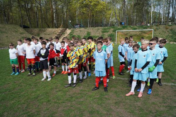 Kilkudziesięciu młodych piłkarzy w białych, czerwonych, żółto-niebieskich i błękitnych strojach stoi na boisku prz