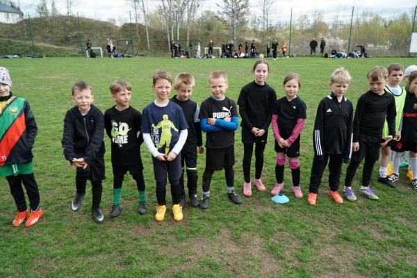 Młodzi piłkarze w sportowych, głównie czarnych strojach stoją w rzędzie na zielonym boisku, a w tle widać kibiców i innych zawodników.